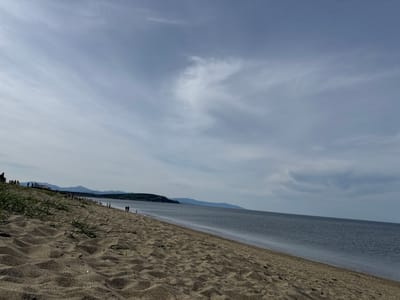 The image shows a beach scene in Okhotskoe, Russia. The foreground is dominated by a sandy beach with sparse vegetation. In the distance, there is a headland and a mountain range. The water is calm and stretches to the horizon. A number of people are visible walking or standing along the beach. The sky is mostly cloudy with some patches of blue.