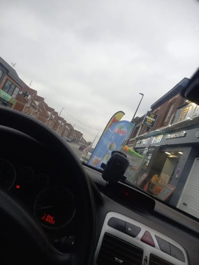 The image depicts a street scene captured from the interior of a vehicle, looking forward through the windshield, on Rue Anatole France in Noyelles-sous-Lens, France. The sky is overcast and grey.
In the foreground, the dark steering wheel of the vehicle occupies the lower-left quadrant. On the dashboard, a red digital display indicates "2940" and "216599," likely trip mileage and total odometer reading. Part of the dashboard, including air vents and a red hazard light button, is visible on the lower-right. A black device mount is attached to the dashboard, centered near the windshield.
Beyond the vehicle, a commercial street is visible. On the right, directly ahead, is a building housing businesses. Signs include "MARKET CITY" with a shopping cart icon, and "SANDWICHERIE." Multiple shopping carts are positioned outside these businesses, along with promotional signs and displays. Some storefronts feature closed metal roller shutters. Two large vertical banner flags are present: one multi-colored (yellow, red, orange) and another blue banner displaying "Oasis."
On the left side of the street, a continuous row of two-story brick buildings with pitched roofs extends into the distance. A partial sign reading "Hobby" is visible on the far left. Streetlights are present, and decorative string lights are strung across the street in the background. Parked vehicles are partially visible along the left side of the road. No individuals are clearly discernible within the scene.