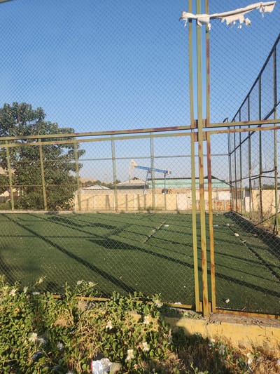 The image shows a fenced-off outdoor area, likely a sports field with artificial turf, under a clear blue sky. In the background, beyond the fence, is a settlement with buildings and vegetation. A prominent feature in the background is a blue and yellow structure resembling an oil pumpjack. The foreground consists of overgrown vegetation and some litter. The overall scene suggests an undeveloped or perhaps abandoned outdoor recreational area.