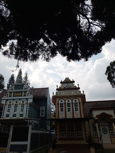 The image displays an exterior architectural scene in Weintiapa, Liberia, showcasing two prominent multi-story buildings under a partly cloudy sky. The building on the left features a dark teal facade with white trim and gold accents on its stepped gable, incorporating multiple white-framed windows. To its right, a tan-colored building presents more elaborate, cream and white ornate gables, also equipped with multiple white-framed windows. An extension of this building visible on the right includes a white entrance door with a circular glass panel. Both structures are topped with reddish-brown tiled roofs. The upper foreground is occupied by a dark silhouette of dense tree branches and foliage. In the background, several tall, slender evergreen trees are visible, extending above the rooftops against the white and blue sky. No individuals, overt actions, or interactions are depicted in the scene.