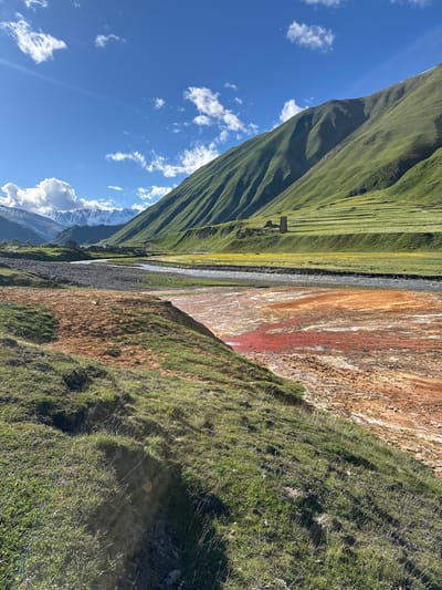 In Ketrisi, Georgia, a natural landscape unfolds under a bright blue sky dotted with white clouds. A winding river cuts through the valley, its water exhibiting vibrant streaks of red and orange, suggesting mineral deposits. On the left bank, a grassy slope leads down to the water, with patches of reddish-brown earth. Across the river, verdant green hills rise steeply, their slopes patterned with terraced fields and a solitary stone tower. In the distance, snow-capped mountains form a majestic backdrop. The scene is devoid of people.