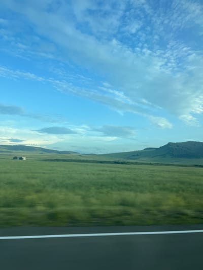The image shows a blurred landscape, likely captured from a moving vehicle on a road. The foreground consists of a dark asphalt road with a white line marking its edge. Beyond the road, a vast, green, grassy field stretches towards rolling hills in the background. The hills are covered in vegetation and appear to be part of a natural landscape. The sky above is a clear blue with scattered, wispy white clouds. In the distance, on the left side of the frame, a small, white building is visible amidst the field. The overall impression is of a scenic, rural environment, possibly in the city of Толстый Мыс, Russia, given the context provided.