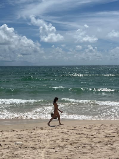 A woman walks along the sandy shore of a beach in Choeng Thale, Thailand. She is wearing a bikini and carrying a bag. The ocean is a deep green with white waves crashing on the shore. The sky is blue with scattered white clouds. The sand is light brown and shows footprints from previous beachgoers. A discarded plastic bottle lies on the sand in the foreground.