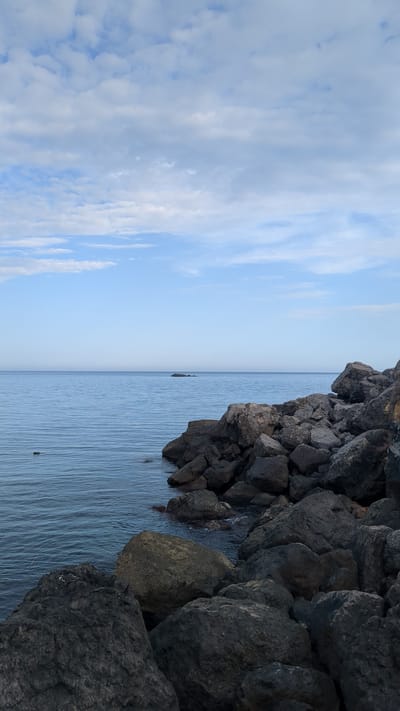 The image depicts a coastline near Bondarenkovo, Ukraine. On the right, dark, jagged rocks form a cliff face that descends into the water. The water is calm with slight ripples. In the distance is an almost indistinguishable horizon line. To the left, the sky is visible with scattered white clouds.