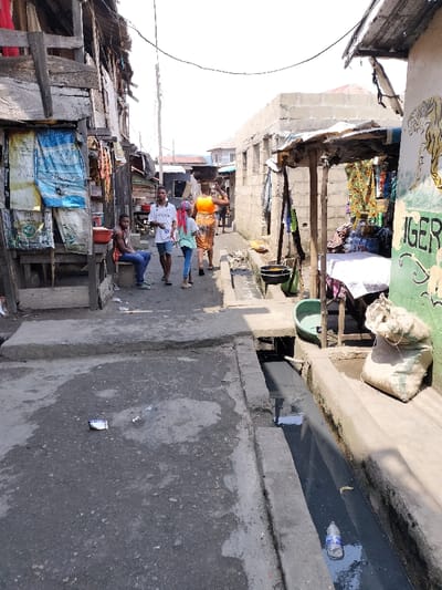 The image depicts a narrow, unpaved street or alleyway in Oworonshoki, Nigeria, under clear skies. The ground consists of uneven, dark-colored concrete and dirt, with scattered refuse, including a white wrapper. An open concrete drainage ditch runs along the right side of the street, containing dark, murky water and a clear plastic bottle.
On the left side, a weathered wooden structure, potentially a stall or building facade, stands. Large, discolored woven sacks, one blue and one white with text, are hung on its front. A male individual, dressed in a red sleeveless shirt and blue jeans, is seated on a low wooden stool next to this structure. A red plastic basin is visible nearby. Further down the left, other informal structures and what appears to be merchandise are present.
On the right side, a partially constructed concrete block building is visible, alongside a makeshift market stall supported by wooden posts and covered by a corrugated metal sheet. Colorful fabrics and other goods are displayed within this stall. A table covered with a white cloth holds additional merchandise. A green plastic basin rests on the ground near the stall, and a light-colored burlap-like sack sits on a concrete ledge bordering the drainage ditch. A wall painted with a yellow tiger and the word "TIGER" in green is also visible on the right.
Four individuals are walking in the middle ground, away from the foreground. The closest, a male, wears a white t-shirt and light shorts, holding a white object. Behind him, a female wearing a light-colored top and dark pants has her hair covered by a pink scarf. Further back, another female, dressed in a bright orange top and a patterned skirt, carries a large, dark object on her head. A fifth, less distinct figure is visible further down the street. Overhead, power lines traverse the scene. The structures on both sides are varied, including wooden planks, corrugated metal, and concrete blocks, reflecting an informal urban environment.