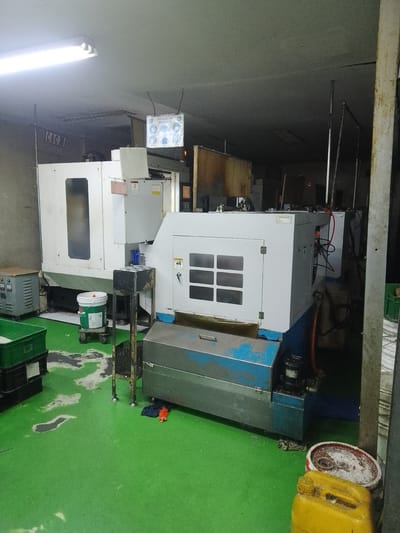 The image depicts a workshop with industrial machinery and materials. In the foreground, a bright green floor is visible, marked with some stains and debris. To the left, green plastic crates are stacked. Towards the center, a large white and blue industrial machine, possibly a CNC machine, dominates the scene. A white bucket with a green label is positioned on a wheeled platform in front of it. Behind this machine, another larger white machine is partially visible.

In the background, a ceiling with fluorescent lighting is present. A sign with "CNC 30" is mounted on a wall on the left. Another display screen showing some images and text is suspended from the ceiling on the right. Various other industrial equipment and materials are scattered throughout the workshop.

The scene suggests an active or recently active industrial environment. The presence of machinery, crates, and buckets points to manufacturing or production activities. The lighting indicates an indoor setting, likely a factory floor or workshop. No people are visible in the image. The overall impression is one of an organized, albeit somewhat cluttered, industrial workspace. The location specified as Ciantra, Indonesia, cannot be visually confirmed from the image itself.