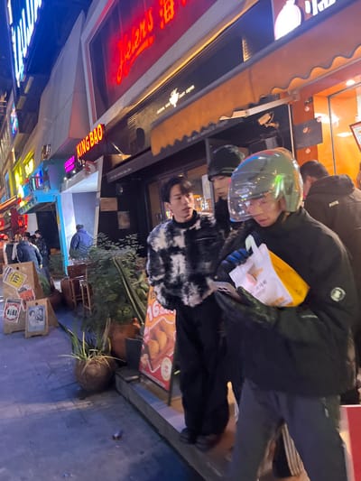 The image, originally oriented vertically but depicting a horizontal street scene, captures a bustling night street in 天宫殿街道, China. Two young individuals are central to the composition. In the foreground, a young man wears a distinctive black and white patterned faux fur jacket, looking directly ahead with a neutral expression. Slightly behind him, another person is dressed in a dark puffer jacket and a reflective, iridescent motorcycle helmet, holding a white paper bag, possibly containing takeout food.
The background is filled with the vibrant glow of various storefronts and neon signs, indicating commercial activity. To the left, a prominent red neon sign spells out "Helen's" in a script font, while another bright pink/purple neon sign reads "KING BAO." A dark sign above a window also displays "CRAFT LIQUOR." A brown, scalloped awning extends over these shops.
To the right of the central figures, a red advertising sign showcases appetizing images of fried food items, resembling nuggets or dumplings, accompanied by the price "16元" (16 Yuan). Potted plants are arranged along the sidewalk, adding a touch of greenery. The ground features scattered objects and smaller signs, some indicating percentages like "89%." The scene is heavily illuminated by artificial lights, creating a lively and energetic urban atmosphere, characteristic of an evening or night setting. People are dressed in jackets, suggesting cool temperatures.