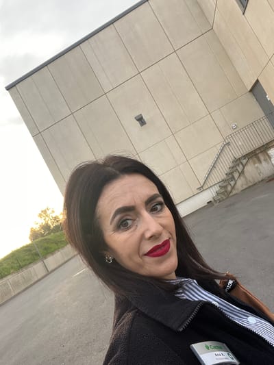 A woman is taking a selfie outdoors. She is wearing dark clothing and a name tag that reads "Cactus Ana A. Poissonnerie". The background features a building with light-colored, rectangular panels, an external metal staircase, a paved area, and some greenery along the ground. The lighting suggests it's near sunrise or sunset. The photograph was taken in Mensdorf, Luxembourg.