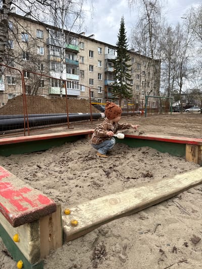A high-angle, slightly Dutch-tilted photograph captures a young child playing in a sandbox in what appears to be an urban setting in Yaroslavl', Russia, under an overcast sky. The child, wearing a brown patterned hat, a plaid shirt, and blue jeans, is squatting in the sandy area, holding a white scoop or shovel in one hand and pointing upwards with the other.
The sandbox is defined by wooden planks, some of which are painted red and green, with yellow caps or details attached. The sand is light brown and appears somewhat coarse, mixed with fallen leaves and debris. To the left of the sandbox, beyond a metal fence, a trench reveals large black pipes, possibly for district heating or water, suggesting ongoing construction or utility work.
In the background, a large, multi-story apartment building with numerous windows and several enclosed balconies dominates the scene. Its facade is a light, somewhat weathered brick or concrete. Bare-branched trees, indicative of late winter or early spring, frame the building and reach into the cloudy sky. A tall, slender conifer stands prominently in front of the building. To the far left, another large, bare-branched tree is visible. No discernible text is visible in the image.