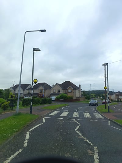 This is a medium-shot, eye-level, vertical photograph of a residential street in Dunfermline, United Kingdom, under an overcast sky. The scene depicts a road with a pedestrian crossing, several houses, streetlights, and a few cars.
In the foreground, the asphalt road is marked with white pedestrian crossing stripes and a broken white line. The left side of the road features a grass verge with some small white wildflowers, bordered by a black metal fence. Several modern residential houses with grey tiled roofs are visible in the background, set behind neatly trimmed hedges. Two prominent streetlights stand tall, one on the left with a simple grey pole and a dark lamp head, and another on the right which is a more complex structure with multiple lamps and a yellow flashing beacon.
In the middle ground, a dark colored car is driving on the road, approaching the pedestrian crossing. Further down the road, two more cars are visible, one red and one white van parked in a driveway. The overall impression is of a quiet suburban neighborhood.
The sky is uniformly grey and cloudy, suggesting a dull or possibly rainy day. The time of day appears to be daytime. There are no people visible in the frame, only the parked and moving vehicles. The setting is clearly residential, with the houses and manicured lawns characteristic of a suburban area. There is a sign on one of the houses indicating a house number or street name, but it is not legible.