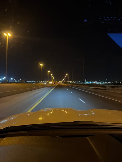 The media file captures a nighttime scene from the interior of a vehicle traveling on a well-lit road in Umm Salal Muhammad, Qatar. The perspective is looking forward from the driver's or front passenger's seat, with the road stretching into the distance.
The multi-lane road features prominent yellow lane markings and is illuminated by a long series of streetlights extending as far as the eye can see, likely positioned along the median and sides of the road. Faint, blurry lights in the distance suggest the presence of other vehicles.
On the left side of the frame, a dark section of the vehicle's dashboard is visible, featuring various illuminated controls and a small screen. Readable text on one button says "ENGINE START/STOP," accompanied by other small symbols such as a clock, a Wi-Fi-like icon, and a car graphic.
The right side of the frame shows a reflection of the vehicle's exterior, which appears to be a light color, possibly white or beige. A windshield wiper and part of the side panel are discernible in this reflection. The overall scene conveys the experience of driving at night on a clear evening, with the extensive artificial lighting providing good visibility of the road ahead.