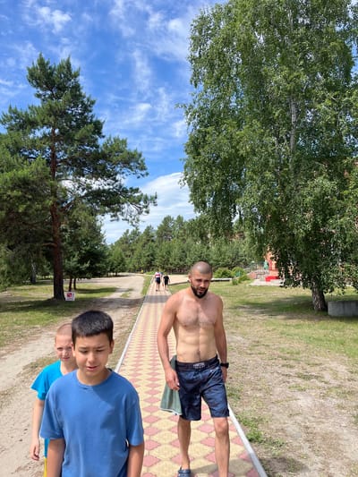 In Lebyazhye, Russia, on a sunny day with scattered clouds, a group of people walk along a paved path bordered by grass and trees. In the foreground, a shirtless man with a beard, wearing dark patterned swim trunks, walks towards the viewer. To his left, two young boys, one in a blue t-shirt and the other in a teal t-shirt, are also walking. In the background, two figures are seen walking further down the path. The environment is characterized by tall evergreen and deciduous trees, with a dirt path running alongside the paved walkway. A red and white sign is visible on a tree to the left of the paved path.