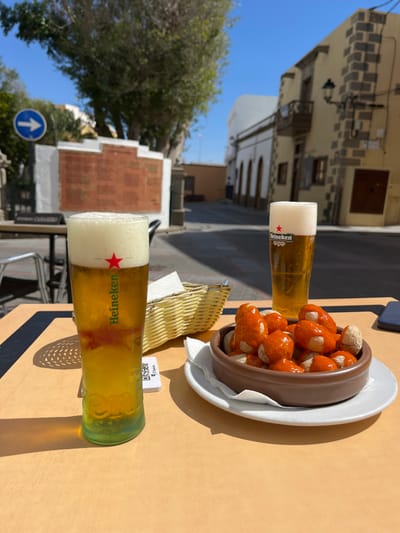 The image shows a table with two glasses of Heineken beer and a bowl of papas arrugadas (wrinkled potatoes) with mojo rojo (red sauce). The scene is a cafe or restaurant patio in the city of Agüimes, Spain, at coordinates 27.907, -15.447. In the background, a white-washed building with a brown tiled roof is visible. The patio is made of concrete, and there is a small tree in the background. The sun is shining brightly, casting shadows from the table and chairs.  The table is made of wood, and it has a brown tablecloth. The glasses of beer are both filled to the brim, and the bowl of papas arrugadas is full. The papas arrugadas are a popular dish in the Canary Islands and are typically served with mojo rojo sauce. This is a scene of casual dining in a sunny outdoor setting.