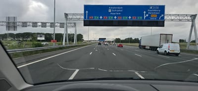 The image provides a driver's perspective from within a vehicle traveling on a multi-lane highway in Hoofddorp, Netherlands, under an overcast sky. The bottom portion of the frame shows the car's dashboard and a cracked windshield.

Ahead, the highway features several vehicles, including a red hatchback, a silver sedan, other cars, a white cargo van with license plate V-93-GJG, and a blue semi-trailer truck with a white trailer.

A prominent blue overhead gantry sign spans the highway, directing traffic. The main part of the sign indicates the route straight ahead for "Amsterdam," "Schiphol," and "Haarlem" via motorways A4 and E19. To the right, an exit sign for "Hoofddorp" and "Aalsmeer" is displayed, designated as exit "3 + 3a" and national road "N 201," with a notice indicating the exit is "600 m" away.

Along the left side of the road, behind a metal guardrail and a line of green trees, are smaller red and white roadside signs reading "UGS ABD ↑" and "UGS C →." Further in the distance, a billboard is visible, depicting what appears to be a car wheel or steering wheel. Streetlights line the highway, and the overall weather appears to be cloudy daytime.