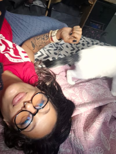 A person is lying on their back on a bed. They are wearing a red t-shirt with white graphic designs, black-rimmed glasses, and have a nose ring. The person has dark hair and visible tattoos on their left arm. A white cat with black markings is present in the image, partially obscuring the person. The bedding includes a blue patterned sheet and a pink quilted blanket with white floral designs. The background shows part of a shelf unit, suggesting an indoor setting. The scene appears to be a personal space, possibly a bedroom. The image does not contain information that places it in Bridlington, United Kingdom.