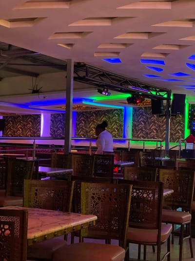 This image captures the interior of a commercial establishment in Kimorori, Kenya, likely a restaurant, club, or event venue, presented from a rotated perspective. The scene is dominated by rows of ornate wooden chairs, featuring intricate, possibly Middle Eastern or African-inspired, geometric and floral carvings. Many chairs have light-colored seat cushions and appear to be either stacked or neatly arranged along tables that are mostly out of frame on the right.
In the middle-ground, a person wearing a white top is visible, bent over near a silver metal railing, seemingly engaged in setting up, cleaning, or arranging the space. Their back is to the viewer.
The venue's decor is notable for its vibrant artificial lighting and textured surfaces. A prominent feature on the left is a light-colored wall with a pattern of geometric cutouts, illuminated from behind by bright blue lights. Above this, a robust metal truss structure supports sound equipment and stage lights, which emit strong green and purple beams. The background walls are adorned with a decorative wallpaper featuring a lush, leafy pattern, further enhanced by strips of blue, green, and purple LED accent lighting. The overall ambiance is lively and suggests an evening or night setting, or an environment designed to be artificially lit. There is no visible text in the image.