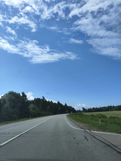 The image shows a two-lane asphalt road in Salacgrīva, Latvia. The road curves to the left and is bordered by a grassy shoulder and a dense line of trees on the left. On the right side of the road, there is a field of green grass with some wildflowers. The sky is bright blue with scattered white clouds. There are two vehicles visible on the road: a red bus in the distance to the left, and a white van further ahead and to the right. Several road markers are visible along the shoulder of the road. The image appears to be taken from a vehicle moving along the road.