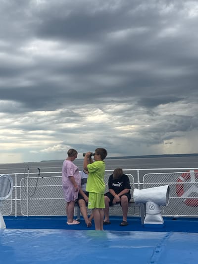 The image depicts a group of people on the deck of a vessel, likely a ferry, near Ikskoe Ust'e, Russia. The sky is overcast with dark, dramatic clouds. In the foreground, the bright blue deck is wet, suggesting recent rain or spray. On the deck, a young boy in a lime green t-shirt and shorts is holding binoculars to his eyes, looking out at the water. Beside him, a woman in a pink t-shirt dress and slip-on shoes stands looking down. To the right, a young person in a black Puma t-shirt and shorts is sitting on a bench, head bowed. A large, silver, horn-shaped object, possibly a speaker or a lookout device, is mounted on a pedestal to the right of the bench. A white railing with a net pattern separates the deck from the water, and a life preserver is visible on the railing. The water is a muted gray, and a distant shoreline with trees is visible under the turbulent sky. The overall atmosphere is somber due to the weather conditions.