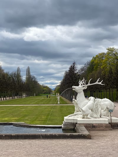 The image shows a statue in the garden of Schwetzingen Palace, Germany. The white statue depicts a man and a deer, with water spouting from the deer's mouth into a pool. The background consists of a manicured lawn, trees, and other statues in the distance, leading to a view of the sky. The sky is overcast with heavy clouds. A narrow canal is positioned close to the viewer.