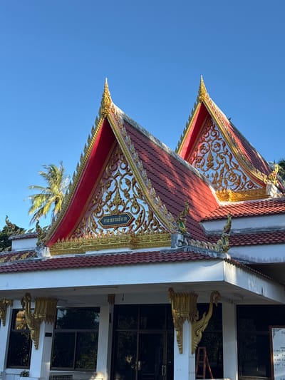 The image shows the exterior of a building with a distinctive multi-tiered roof structure in Baan Tha Po, Thailand. The roof is adorned with red tiles and intricate golden ornamentation, including decorative patterns and finials. The building's facade features large, dark-tinted windows, and ornate golden brackets are mounted on white pillars supporting the roof overhang. A blue sky serves as the backdrop, with the fronds of a palm tree visible in the mid-ground. The scene is well-lit, suggesting it was captured during daylight hours. There are no people visible in the image. The architectural style appears to be of a temple or a similar religious or cultural building.