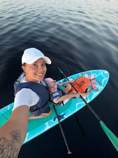 A woman and a child are on a paddleboard on a body of water. The woman is wearing a white cap, a white t-shirt, and a blue life vest. She is smiling and looking at the camera. The child is wearing a pink hat and a blue life vest. The child is holding a paddle. The paddleboard is decorated with a blue and white pattern and has an orange waterproof bag and two pairs of flip-flops on it. The water appears dark and calm, suggesting it might be late in the day. The scene takes place in Salaspils, Latvia.