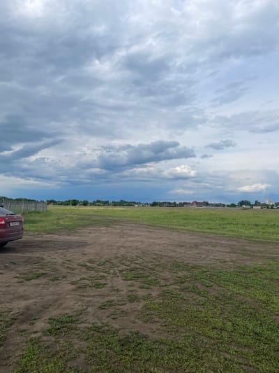 The image depicts a view of Нижнее Санчелеево, Russia. In the foreground is a field of grass and dirt, with a red car partially visible in the lower right corner. A fence line borders the field. Beyond the field is a line of trees and buildings, suggesting the edge of the village. The sky above is filled with gray and white clouds.