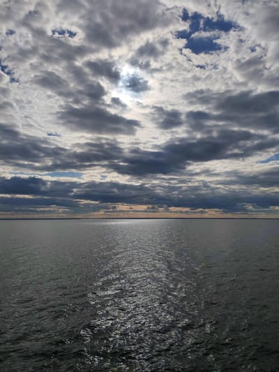 The image displays a vast natural landscape, primarily composed of a large body of water extending to a distant horizon under a dramatic, cloudy sky. The setting is outdoors, likely a large lake or sea near Sannikovo, Russia.
The upper portion of the frame is dominated by a dense array of cumulus and stratocumulus clouds, varying in shades from bright white where sunlight hits them, to dark grey and blue-grey. Patches of clear blue sky are visible through openings in the cloud cover, particularly towards the left and upper right. The sun, partially obscured by clouds in the upper-middle, creates a bright, diffused glow that filters through and casts a strong, shimmering reflection across the water.
The water surface in the foreground and midground is textured with numerous small ripples and waves, catching the sunlight to form a sparkling, elongated path that stretches from the horizon towards the viewer. The water appears dark grey or deep green, reflecting the overcast conditions. A thin, dark, almost imperceptible line along the distant horizon suggests a far-off landmass or coastline.
No people or man-made objects are visible. The time of day appears to be daytime, possibly late morning or afternoon, indicated by the quality and angle of the sun's reflection. The weather is partly cloudy to heavily overcast, with the sun breaking through intermittently. The scene evokes a sense of vastness and natural tranquility. No text is visible in the image.