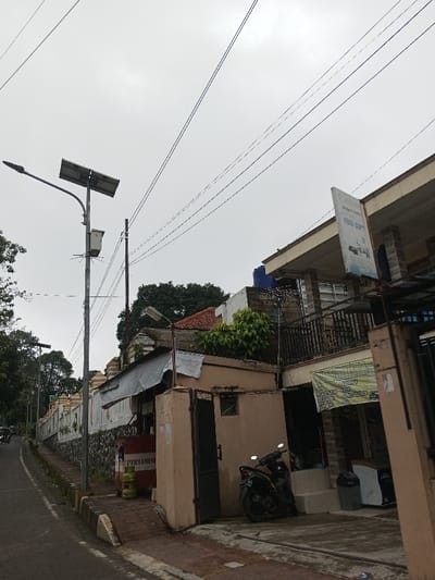 The image displays an urban street scene under an overcast sky. On the left, an asphalt road curves gently downhill, bordered by a concrete curb and a sidewalk. A tall metal streetlamp, equipped with a solar panel, a gray utility box, and wire protrusions, stands prominently on the sidewalk. Behind it, a thinner utility pole supports multiple overhead electrical wires that extend across the upper portion of the frame. A long stone wall, topped with a decorative white and yellow section, runs along the left side of the sidewalk.
On the right side of the street, a series of buildings are present. The foreground features a two-story building with a light-colored facade, a dark-railed balcony, and a vertical blue and white sign displaying the text "FOTO COPY." Below this, a banner with faded text, partially reading "JUAL PULSA," is affixed. Adjacent is a single-story building with a beige exterior and a dark, open metal gate, featuring a corrugated metal awning. In front of this building, a red stand branded "PERTAMINA" holds two green LPG gas cylinders on the sidewalk. A dark scooter, with a visible license plate ending in "3892," is parked on the sidewalk near two dark gray trash receptacles. A smaller outdoor light fixture is attached to a pole near the single-story building. Green foliage is visible on rooftops and in the background. No people are present. This scene is located in the city of Ambakirano, Madagascar.
