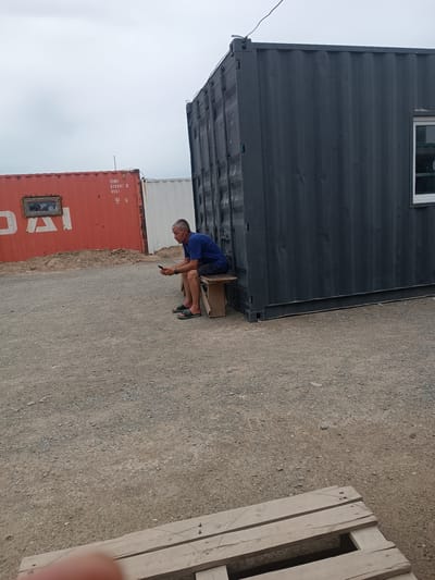 A man sits on a wooden bench outside of a large, dark grey shipping container. He is wearing a blue t-shirt and black shorts and is looking at his phone. To his left is a red shipping container with a window. The ground is covered in gravel and dirt. A wooden pallet is visible in the foreground. The sky is overcast.