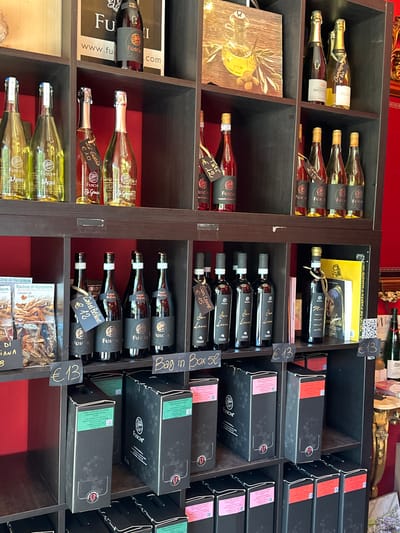 The image shows shelves in a shop displaying bottles of wine and boxes of wine. The shop is located in Silvi, Italy. On the top shelf, there are bottles of white wine and red wine. Some of the bottles have tags with prices. The middle shelf also contains bottles of wine. The bottom shelves have boxes of wine, some with price tags. The walls of the shop are red. The overall scene is well-lit and organized.