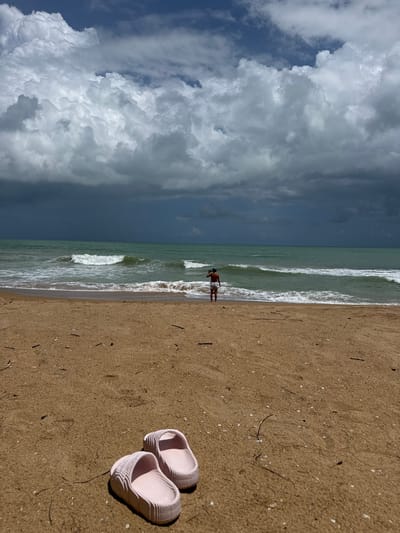 In Ban Khuk Khak, Thailand, a person stands near the shoreline of a beach. The sand is a light brown, with scattered debris such as small twigs and shells. In the foreground, a pair of light pink slide sandals rests on the sand. The ocean is a greenish-blue, with moderate waves breaking on the shore. The sky is dramatic, dominated by large, dark, and textured storm clouds, with patches of blue sky visible. The person, facing away from the camera, is wearing white shorts and a dark top.