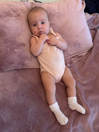 A full shot from an elevated angle shows a baby lying on a plush pink surface, likely a bed or blanket, next to a large pink pillow. The baby is facing upwards, with their head slightly tilted and their hands clasped near their chin. They are wearing a light pink onesie with a subtle textured pattern and white socks. The background is a soft, diffused pink, creating a warm and gentle atmosphere. The scene appears to be indoors, in a residential setting within Wolfsburg, Germany.