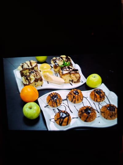 A top-down, slightly angled view of a dessert platter featuring muffins, cake slices, and fruit. The platter is white and irregularly shaped, with distinct sections. One section holds several small, round muffins topped with dark chocolate drizzle. Another section contains cake pieces, also drizzled with chocolate, along with slices of banana and some mint leaves. A long, pale yellow banana slice is positioned between the cake and muffin sections.
The platter is placed on a dark, flat surface, possibly a table, with a black background. To the left of the platter, there is a bright green apple. Below the platter, towards the right side of the frame, is an orange and another green apple. The overall presentation suggests a dessert being served, with a focus on sweet baked goods and fresh fruit. The lighting is somewhat dim, creating a dramatic contrast between the brightly lit food and the dark surroundings. There is no visible text in the image. The scene appears to be indoors, possibly a restaurant or home setting.