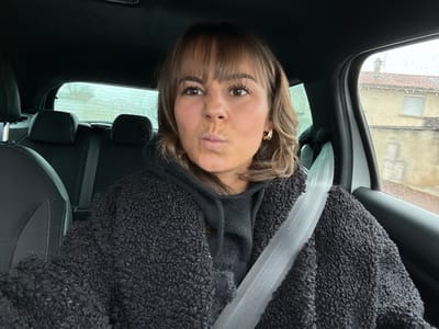 The image displays an adult female in the driver's seat of a vehicle in Saint-Cyprien, France, during daylight hours with apparent rain outside. The female has medium-length brown hair with bangs and is wearing gold-colored hoop earrings. Her facial expression features pursed lips and squinted eyes. She is dressed in a black hooded sweatshirt underneath a textured black jacket. A light-colored seatbelt is fastened across her chest and right shoulder. The vehicle's interior is dark, with black upholstered seats visible, including the driver's seat and two rear passenger seats with headrests. Raindrops are visible on the vehicle windows. Through the front passenger window, a building with light-colored walls and a red-tiled roof is discernible. The view through the rear window is blurred.
