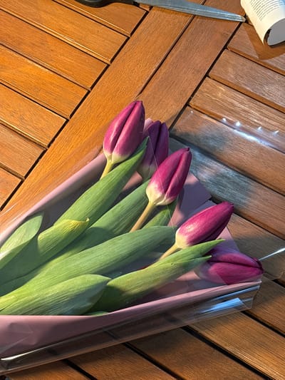 A bouquet of five purple tulips, with vibrant green stems and leaves, is centrally positioned on a wooden slatted surface, likely a table. The tulips are wrapped in a translucent plastic film over a light purple paper backing. Sunlight or strong artificial light illuminates the scene from the upper left, casting distinct shadows and highlighting the texture of the wood and the tulips. In the upper left background, partially visible, are a pair of silver metal scissors with dark handles, resting on the wooden surface. To the upper right, a piece of white crumpled paper with visible Russian text is also present. No individuals are visible. The scene is located in Trudobelikovskiy, Russia.