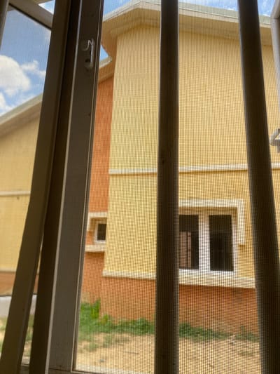 The image shows a view through a window screen towards a yellow building with orange trim. The building has a small, dark-paned window visible. Green foliage can be seen to the right of the building. The screen itself is a mesh, with horizontal bars of the window frame partially obscuring the view. The sky, with some clouds, is visible in the lower left portion of the image, suggesting an outdoor scene. The overall impression is one of looking out from an interior space. The time of day appears to be daytime, given the bright light. There are no people or specific activities depicted, and no visible text. The location context is Bulamare, Nigeria.
