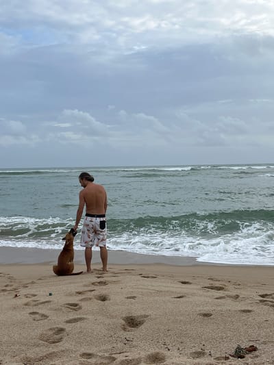 The media file shows a man and a dog on a beach. The man is shirtless, wearing patterned swim trunks, and has his hair tied back. He is standing facing the ocean, with his arm extended towards the dog. The dog, a tan-colored breed, is looking up at the man, with its head tilted. The dog appears to be interacting with the man, possibly being petted or played with. The scene is set on a sandy beach with footprints visible in the sand. The ocean is in the background, with waves breaking on the shore. The sky is overcast with clouds, suggesting it might be a cloudy day. The image is oriented vertically, and the camera is positioned at a medium distance from the subjects. The overall mood appears to be relaxed and casual, suggesting a leisurely day at the beach.