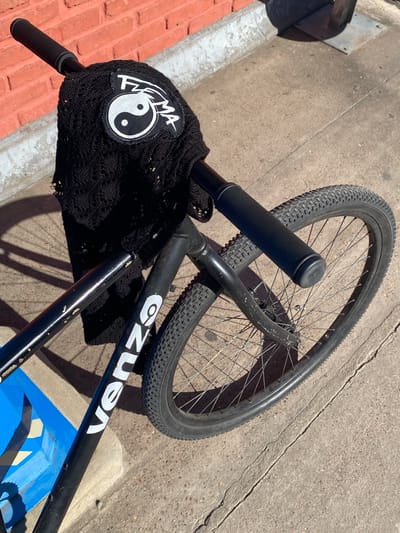 This image presents a close-up, high-angle view of a black bicycle resting on concrete pavement under bright sunlight, likely in Luis Guillón, Argentina. The bicycle is angled across the frame, with its front wheel and handlebars prominently featured.

The bicycle has a black frame, with a partially visible white text logo "VEnz" (possibly "Venza" or similar) on its downtube. Black, textured handlebar grips are visible, and a loosely knitted black cover or bag is draped over the handlebars. Attached to this cover is a white patch displaying a yin-yang symbol and the text "FEMA". The bicycle's front wheel, fitted with a knobby tire, is also clearly visible.

The setting appears to be an outdoor urban or suburban environment. The ground consists of rough concrete with visible cracks and imperfections. A red brick curb runs along the left side of the frame, bordering the concrete. A small, blue object, possibly part of a bin or a painted surface, is visible beneath the bicycle's frame in the lower right portion of the image.

The sharp shadows cast by the bicycle and its components indicate a sunny day, suggesting the time is around midday or early afternoon. No people are present, and the bicycle seems to be momentarily parked or placed aside, implying a pause in its use rather than active riding.