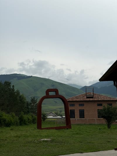 The image is a medium shot of an outdoor scene with a large, dark red, bell-shaped sculpture in the foreground. The sculpture is made of metal and appears to be an abstract representation of a bell. Behind the sculpture, a two-story brick building with a red tile roof is visible. The building has several dark windows and a ladder-like structure on its roof. To the left of the building, a dense line of green trees stands. In the background, rolling green hills covered in grass and trees rise up to meet a cloudy sky. The overall environment suggests a rural or mountainous location. This scene is located in Shabdan, Kyrgyzstan.