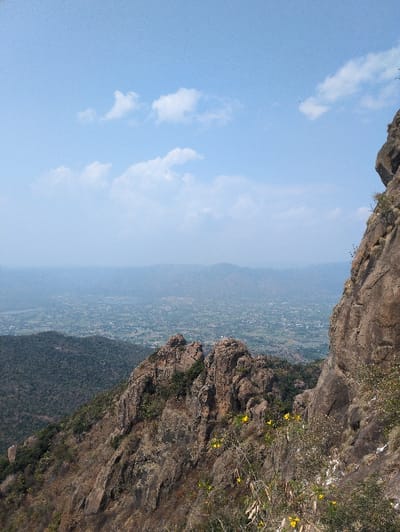 The image displays an elevated landscape view overlooking the region of Kadaladi, India. In the immediate foreground to the right and center, a steep, rocky mountain slope is visible, characterized by prominent brown and grey rock outcrops. Sparse, dry scrub vegetation and several clusters of small yellow flowers grow among these rocks. The midground features lower hills covered in dense, dark green foliage, sloping down towards a valley. The background reveals a wide, hazy valley floor populated with numerous small structures and cultivated fields, indicating settlements and agricultural activity. The sky above is light blue with scattered, diffuse white clouds. No individuals or specific actions are depicted.