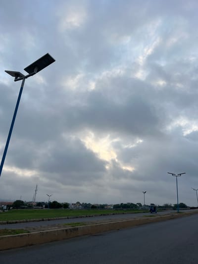 The image shows an overcast sky above Abeere, Nigeria, with a view along a paved roadside. Modern blue streetlights are spaced along the roadside, which has a strip of green grass separating it from the buildings in the background. A single blue three-wheeled vehicle, possibly an auto rickshaw, is visible on the road. The sky is predominantly gray, with a lighter area where the sun is breaking through the clouds.