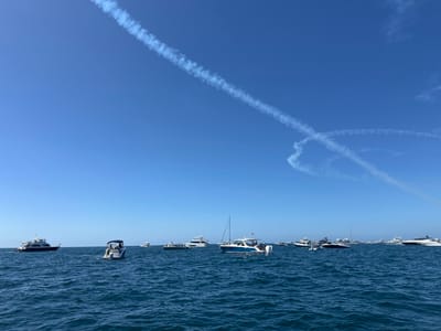 This image displays the coastal waters off Huntington Beach, United States, under a clear blue sky. White vapor trails from an aircraft are visible in the upper atmosphere, forming an "X" shape towards the right and a distinct diagonal line extending from the upper left. The ocean surface is a deep blue with moderate choppiness. Numerous recreational boats are distributed across the water, primarily consisting of motorboats and yachts. A white motorboat with a dark blue hull stripe and a small mast is positioned in the mid-left. Centrally, a smaller white motorboat with a dark canopy is visible, oriented with its stern facing the viewer, containing several occupants. Additional white and dark-hulled vessels of varying sizes are stationary or slowly drifting throughout the midground and background, indicating a gathering of marine traffic. No specific actions or interactions beyond passive observation are discernable among the people on the boats.