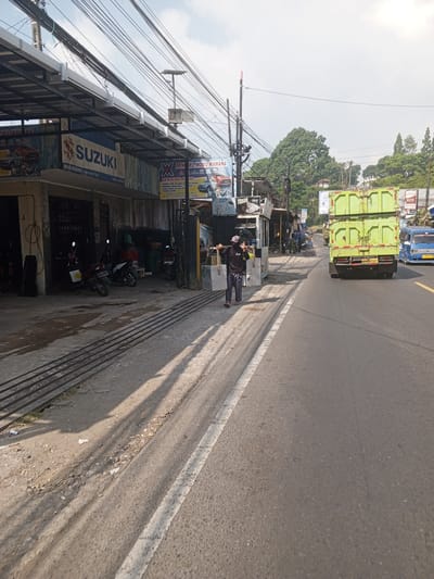 The image depicts a street scene. A person wearing a dark shirt, face mask, and blue jeans is walking on the side of a road. They are carrying two long poles over their shoulders. To the left of the road, there is a row of buildings with shops, including one with a "SUZUKI" sign. Several motorcycles are parked in front of the shops. A large green dump truck is driving on the right side of the road, and a blue van is behind it. The road is paved and has white lane markings. Overhead, there are numerous electrical wires. The environment appears to be urban or semi-urban, with trees visible in the background and a cloudy sky. The image does not contain any elements that would suggest the location is Ruwan Eliya, Sri Lanka.