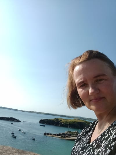 A woman is taking a selfie with a bright blue sky and a coastal landscape in the background. She has short, blonde hair and is wearing a black and white patterned dress. The background shows the sea with several small boats scattered across the water. Rocky islands with green vegetation dot the sea. In the distance, a hazy landmass can be seen. A concrete or stone wall is visible in the foreground.