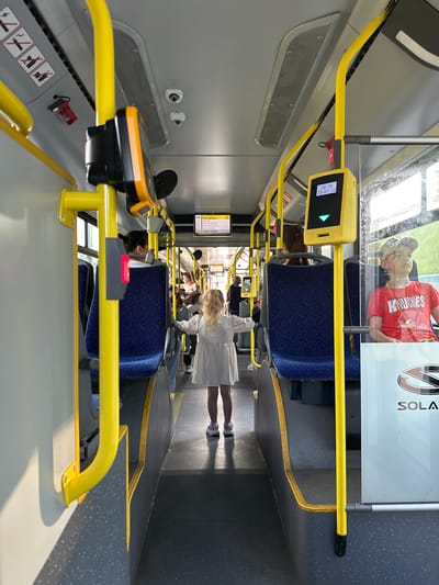 The image shows the interior of a bus in Sosnowiec, Poland, specifically on a Solaris bus, as indicated by the logo on the right. The aisle of the bus is visible, with yellow handrails and blue seats. In the foreground, a young girl with blonde hair in a white dress stands with her arms outstretched, facing away from the camera. Further down the aisle, several passengers are seated, including a woman with dark hair and a man wearing a red t-shirt and a baseball cap. A ticket validation machine is visible on the right, displaying the time and date "18 29 13-07-26". The lighting suggests it is daytime.