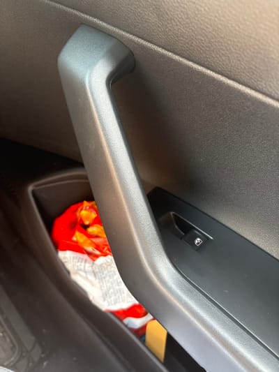 The image shows the inside of a car door. The door panel is black and textured. A black plastic door handle is visible. There is a storage compartment in the door panel, containing a red and white bag of what appears to be snacks.  The door panel also has a window control switch, which is black and has a silver symbol of a window. The coordinates provided place the location in a residential area of San Rafael, Costa Rica.