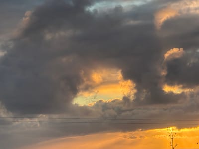 Golden light peeks through the turbulent, gray clouds filling the sky. A small opening reveals a glimpse of the setting sun. Thin branches hang in the upper left corner, silhouetted against the bright light. Two electrical wires stretch horizontally across the image. The clouds are dense and layered, creating a dramatic and atmospheric scene.
