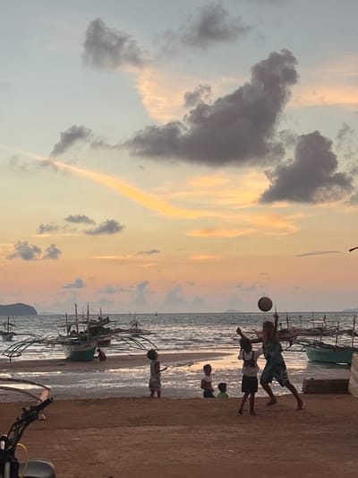 The image depicts a sunset scene at a beach in Cuyo, Philippines. In the foreground, a sandy beach is visible, with the handlebars of a motorcycle on the left. Further back, the ocean meets the shore, with several traditional outrigger boats partially submerged in shallow water. A group of children are playing on the beach and in the water. Two children are actively engaged in a game of volleyball, with one jumping to hit the ball in the air. Other children are observing or playing nearby. The sky is filled with dramatic clouds in shades of orange, yellow, and gray, indicating the setting sun. The overall atmosphere is one of a peaceful evening by the sea.