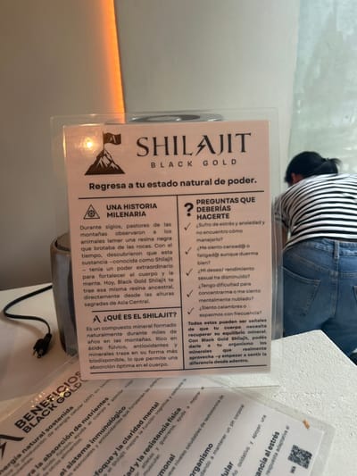 The image shows a sign advertising a product called "Shilajit Black Gold" at a store in Pozos, Costa Rica. The store is located at the coordinates 9.952, -84.196. The sign lists the benefits of the product, which include boosting energy levels, supporting the immune system, promoting mental clarity, and aiding physical performance. It also includes a list of questions to ask oneself to determine if Shilajit is needed. A person is reading the sign, bent over, as if to get a closer look. Their lower back, right leg, and foot are visible, as well as their right arm and the upper part of their head. The sign is in a glass case, behind which is a beige wall. The environment is dimly lit, and a faint orange glow is visible in the top left corner of the image.