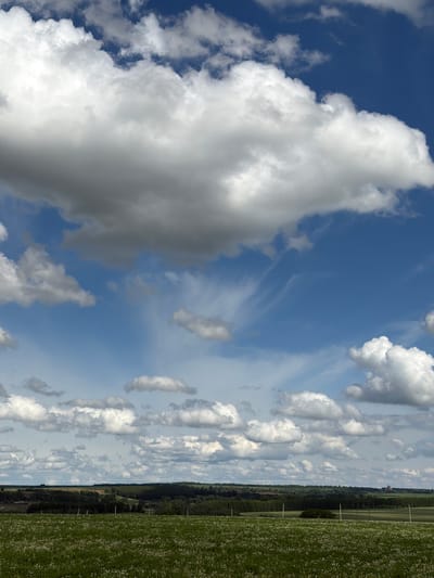 The image shows a bright, partly cloudy sky over Udmurtskoe Kizekovo, Russia. Large, white cumulus clouds occupy the left side of the frame against a deep blue sky. Towards the right, thinner, more scattered clouds stretch across a lighter blue expanse. A thin strip of land with trees and a field lies along the right edge of the image.